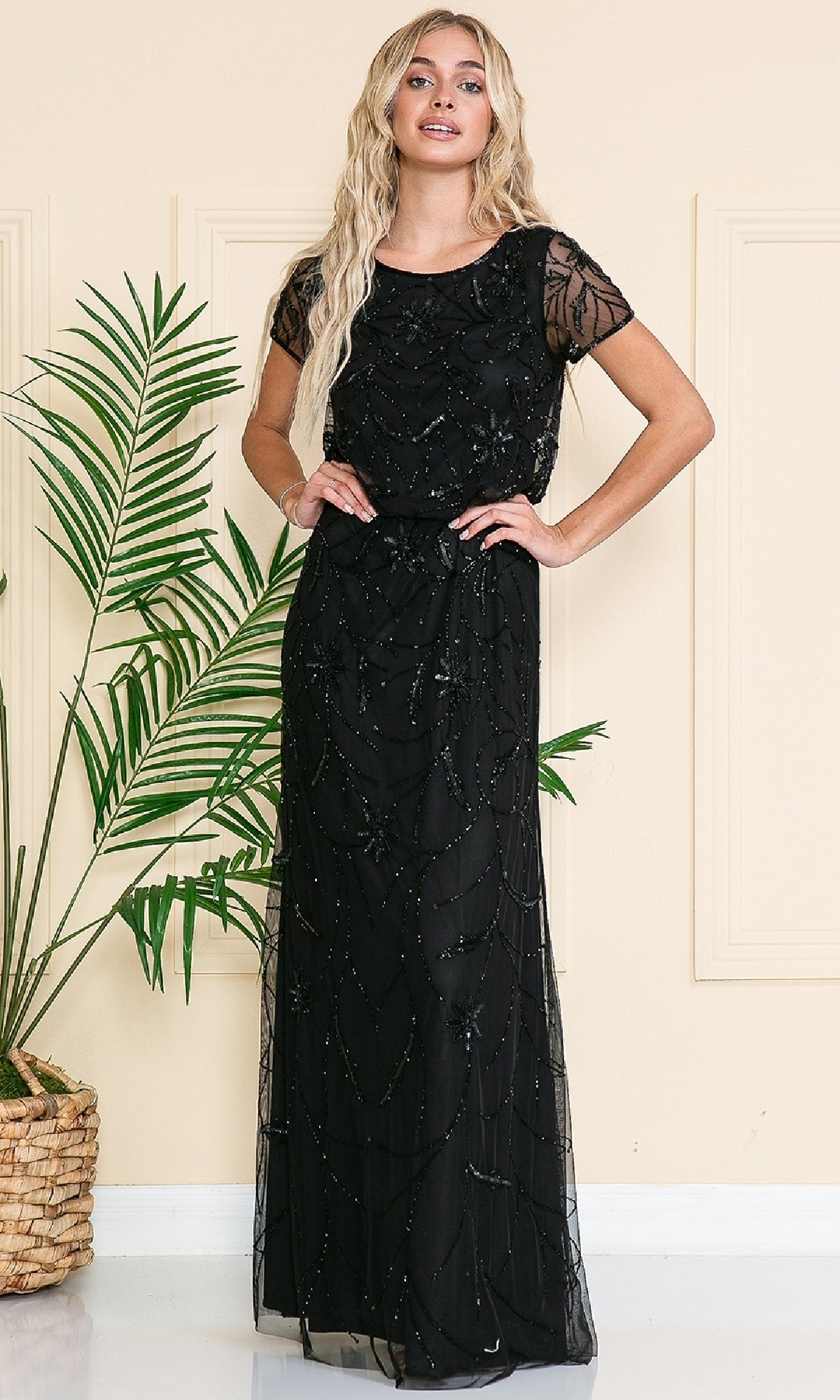A woman stands indoors in the Blouson Long Formal Dress: Amelia Couture IN004, a floor-length black gown with sheer short sleeves and beaded details, next to a green potted plant.